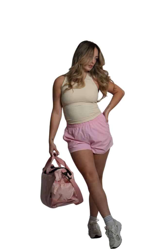 A woman with long, wavy blonde hair wears a beige sleeveless top, pink shorts, white sneakers and socks. She carries the TRIARC Duffle Bag in pink and stands against a white background.