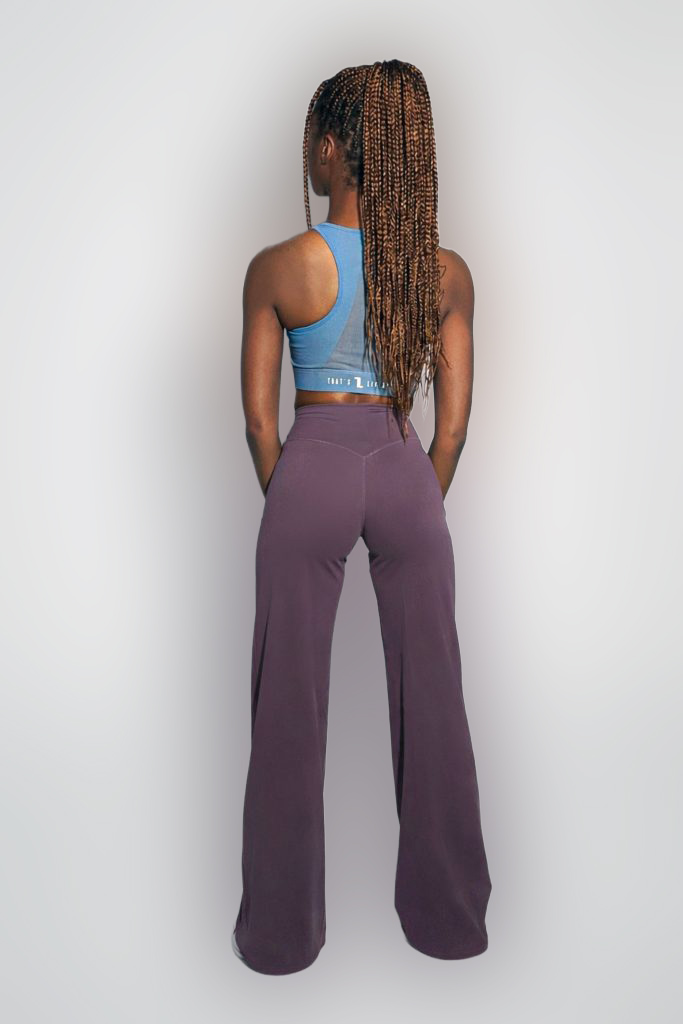A person with long, braided hair stands facing away, wearing a blue crop top and high-waisted, wide-leg purple pants against a light gray background.