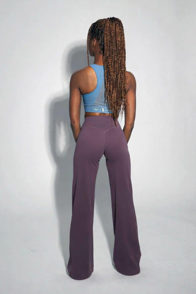 A person with long, braided hair stands facing away, wearing a blue crop top and high-waisted, wide-leg purple pants against a light gray background.