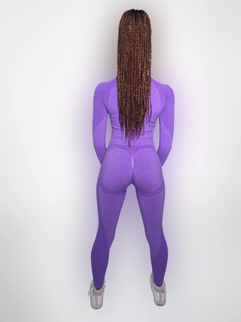 A person with long braids is standing facing away from the camera, wearing a fitted purple athletic outfit and white sneakers, against a plain white background.