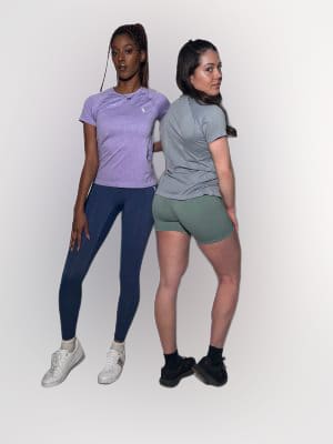 Two women pose in athletic wear against a plain white background. One wears a purple shirt with blue leggings and white sneakers, the other wears a gray shirt with green shorts and black sneakers.