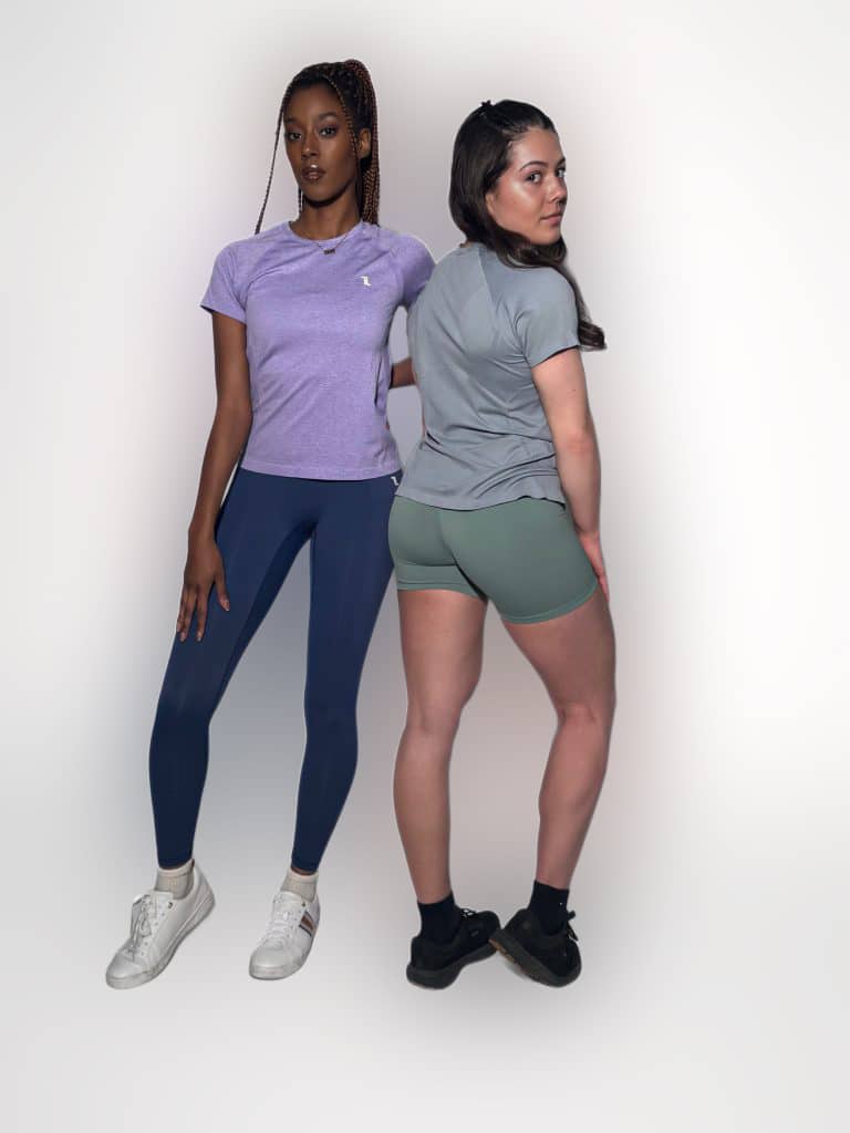 Two women pose in athletic wear against a plain white background. One wears a purple shirt with blue leggings and white sneakers, the other wears a gray shirt with green shorts and black sneakers.
