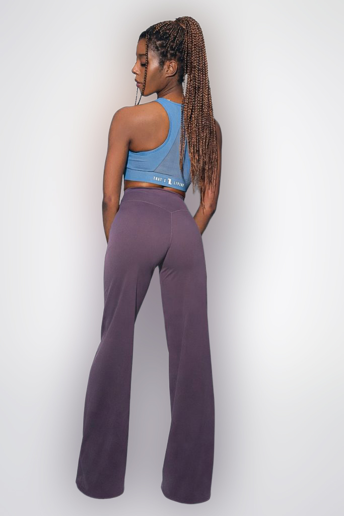 A woman with long, braided hair stands facing away, wearing a blue sports bra and high-waisted, wide-leg purple pants against a light gray background.
