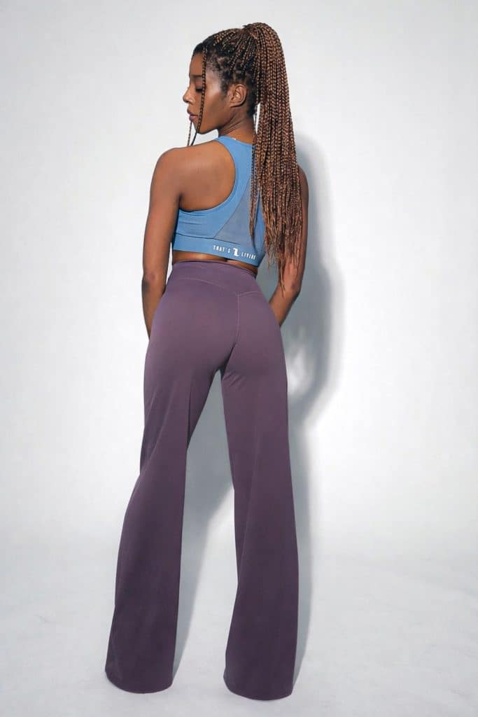 A woman with long, braided hair stands facing away, wearing a blue sports bra and high-waisted, wide-leg purple pants against a light gray background.