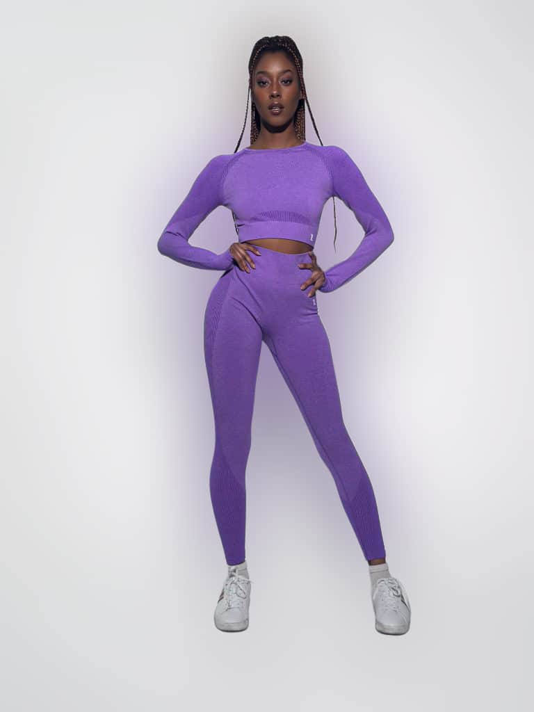 A woman with long braided hair stands confidently against a white background, wearing the Allure Set V2—a matching purple long-sleeve crop top and leggings—paired with white sneakers and her hands on her hips.