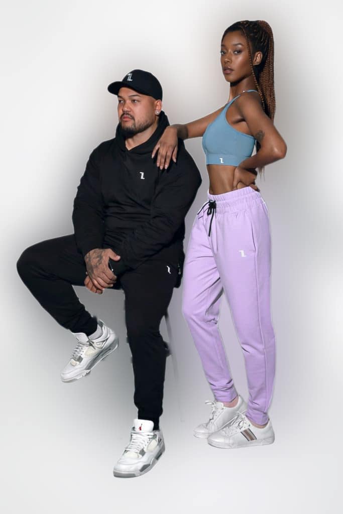 A man in a black tracksuit and cap sits on a stool, while a woman in a blue sports bra and lavender joggers stands beside him with one arm on his shoulder. Both wear white sneakers against a plain background.