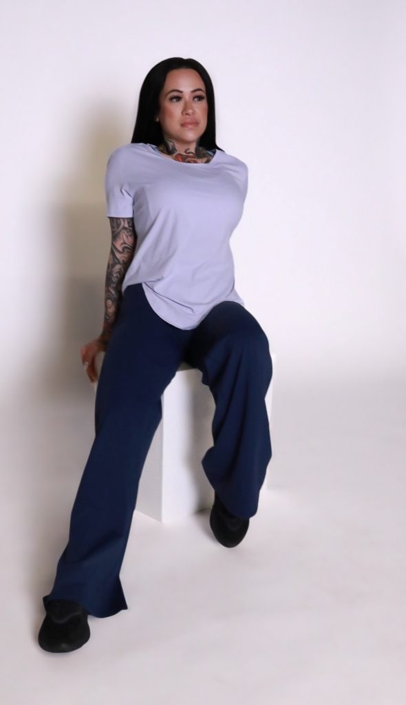 A person with long dark hair, visible tattoos, and dark wide-leg pants wears the VeloraFlow Tee while sitting on a white rectangular block against a plain white background.