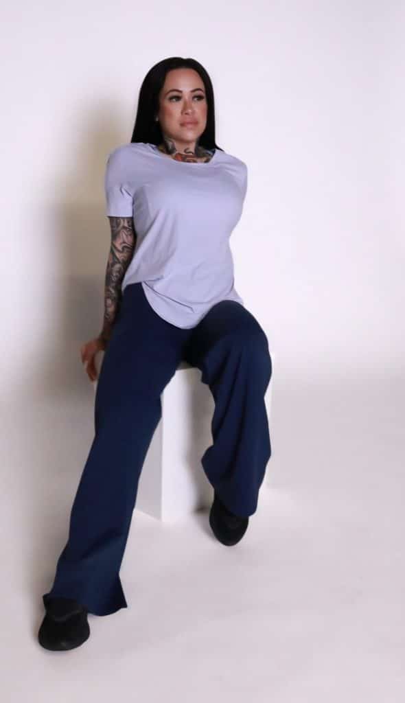 A person with long dark hair, visible tattoos, and dark wide-leg pants wears the VeloraFlow Tee while sitting on a white rectangular block against a plain white background.
