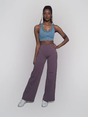 A woman poses confidently against a white background, wearing a light blue zip-up sports bra, high-waisted wide-leg purple pants, and white sneakers. She has long braids and a tattoo on her upper left arm.
