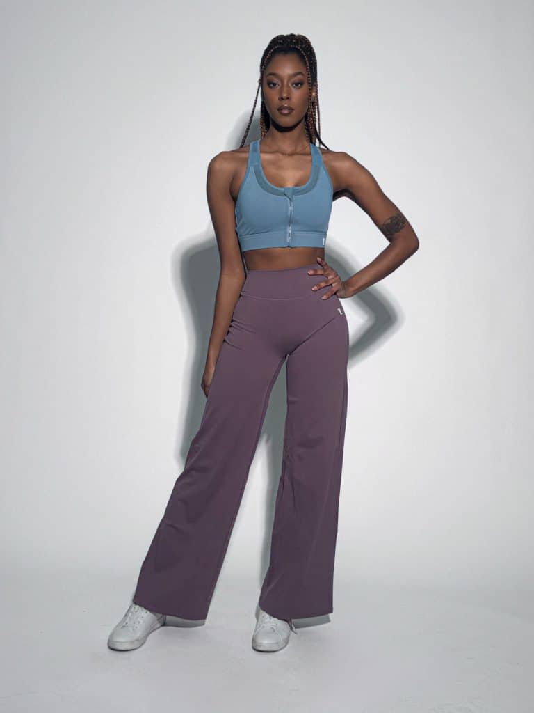 A woman poses confidently against a white background, wearing a light blue zip-up sports bra, high-waisted wide-leg purple pants, and white sneakers. She has long braids and a tattoo on her upper left arm.