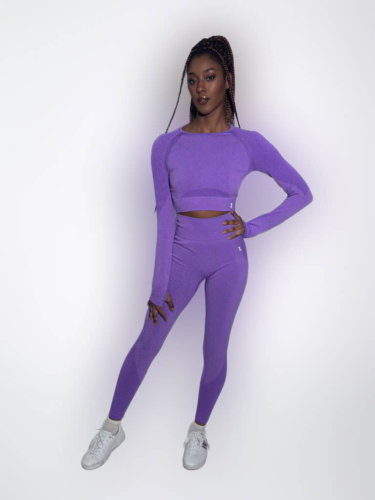 A woman poses confidently against a plain white background, wearing a matching purple long-sleeve crop top and leggings set, with white sneakers.