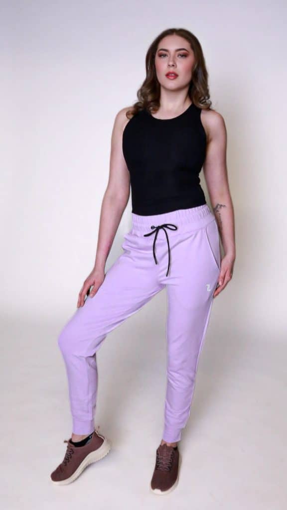 A person poses against a plain background, wearing a black sleeveless top, light purple jogger pants with a drawstring, and brown sneakers. They have wavy hair and a neutral expression.
