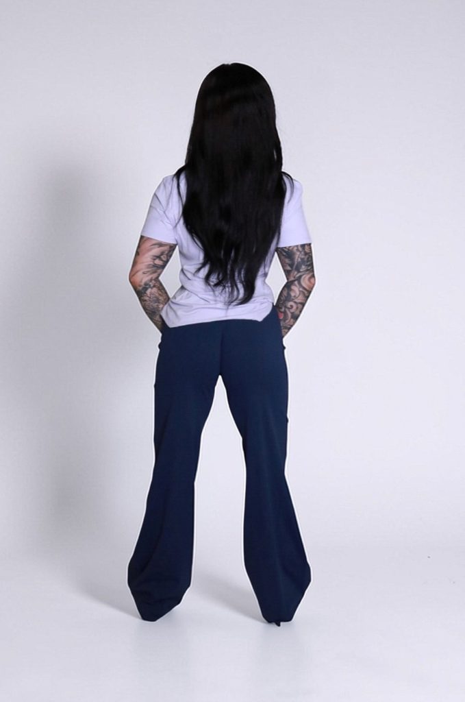 A person with long dark hair and arm tattoos, wearing the VeloraFlow Tee and dark blue pants, stands facing away from the camera against a plain white background.