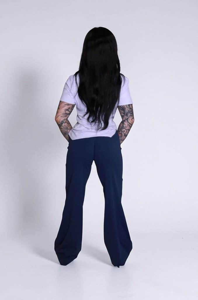 A person with long dark hair and arm tattoos, wearing the VeloraFlow Tee and dark blue pants, stands facing away from the camera against a plain white background.