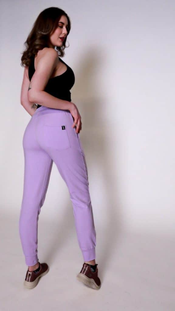 A person with long wavy hair is standing sideways, wearing a black sleeveless top, lavender jogger pants, and brown sneakers against a plain white background.