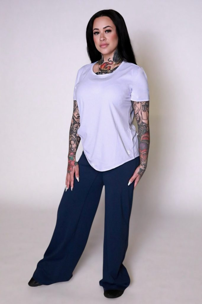 A woman with long dark hair and visible tattoos confidently wears the VeloraFlow Tee, paired with loose navy blue pants and black shoes, standing against a plain light background.