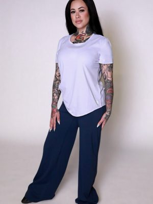 A woman with long dark hair and visible tattoos confidently wears the VeloraFlow Tee, paired with loose navy blue pants and black shoes, standing against a plain light background.