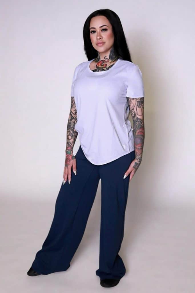 A woman with long dark hair and visible tattoos confidently wears the VeloraFlow Tee, paired with loose navy blue pants and black shoes, standing against a plain light background.