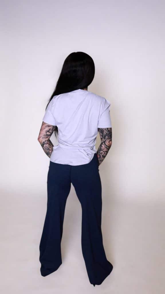 A person with long dark hair, visible arm tattoos, and dark wide-legged pants wears the VeloraFlow Tee in light gray, standing with their back to a plain light background.