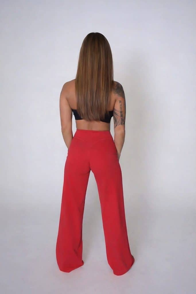 A woman with long straight hair, wearing a black crop top and high-waisted red pants, stands facing away from the camera against a plain light background.