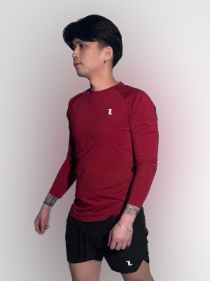 A person with dark hair, wearing a long-sleeved maroon athletic shirt and black shorts, stands against a plain light background. They have visible tattoos on their arms and a slight shadow is cast on the wall.
