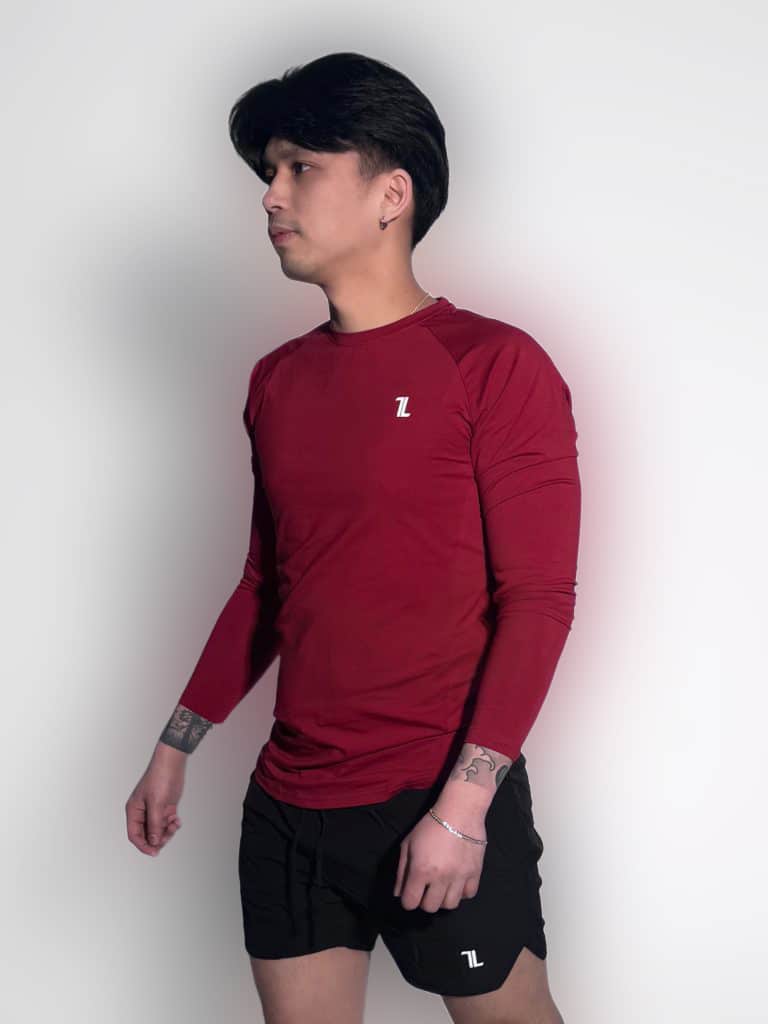 A person with dark hair, wearing a long-sleeved maroon athletic shirt and black shorts, stands against a plain light background. They have visible tattoos on their arms and a slight shadow is cast on the wall.