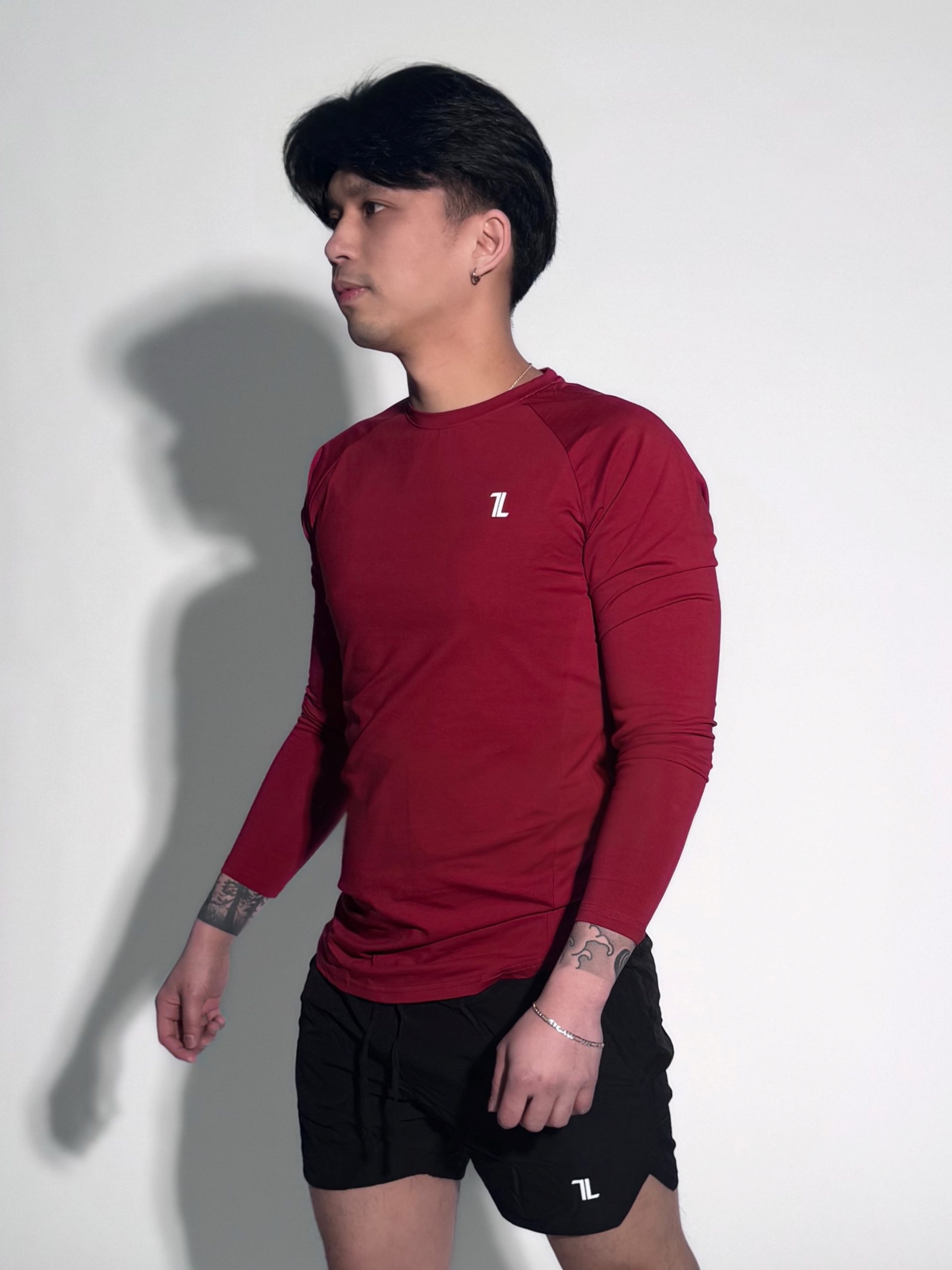 A person with dark hair, wearing a long-sleeved maroon athletic shirt and black shorts, stands against a plain light background. They have visible tattoos on their arms and a slight shadow is cast on the wall.