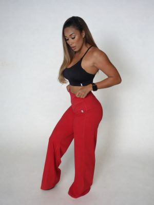 Woman standing against a plain background, wearing a black crop top and wide-legged red pants, looking down with one hand on her hip.