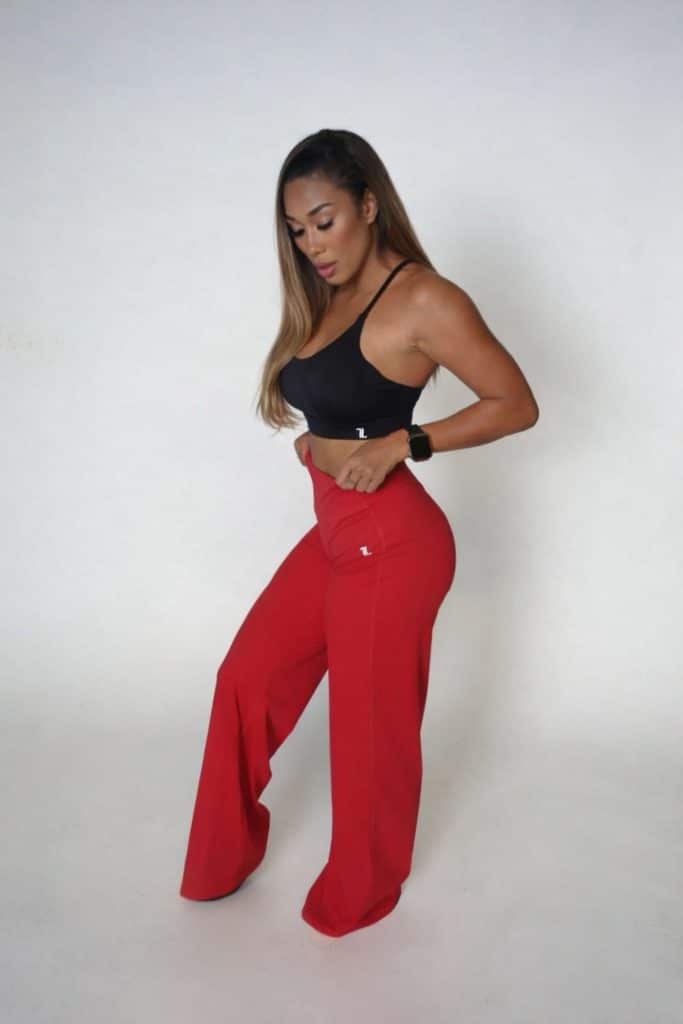 Woman standing against a plain background, wearing a black crop top and wide-legged red pants, looking down with one hand on her hip.
