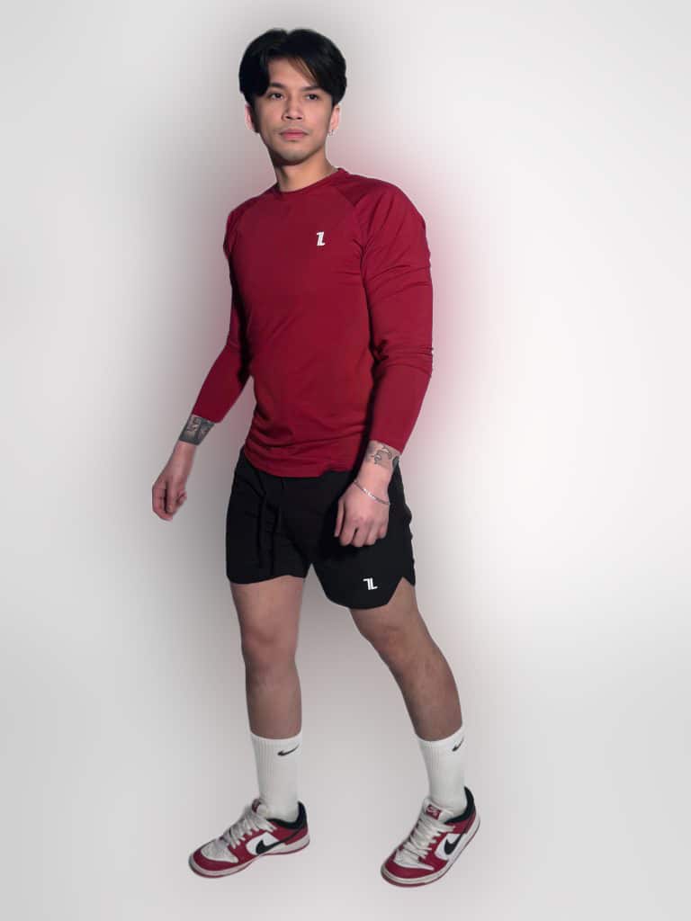 A man wearing a red long-sleeve athletic shirt, black shorts, white knee-high socks, and red-and-white sneakers stands against a plain white background, looking slightly to his left.