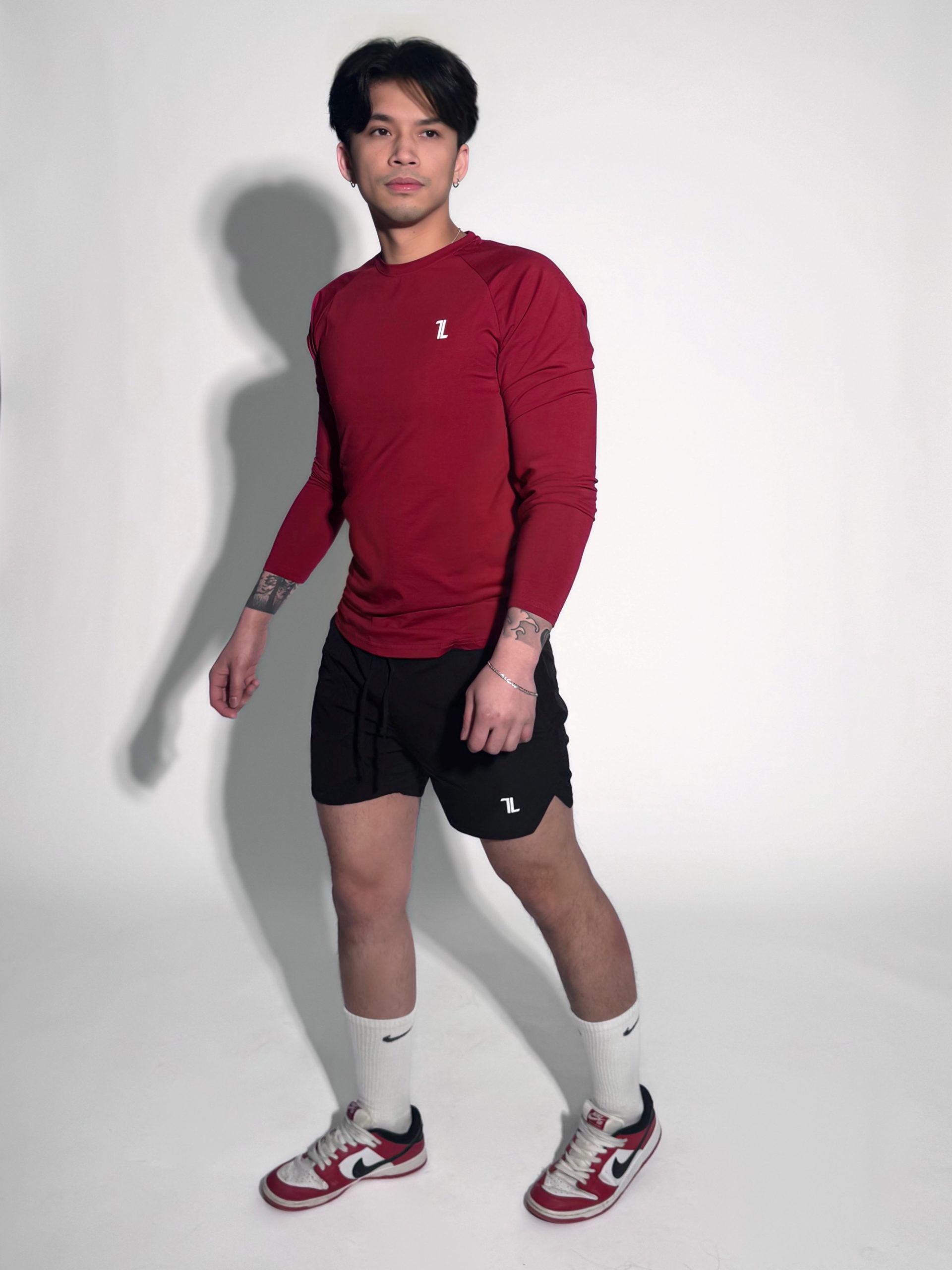 A man wearing a red long-sleeve athletic shirt, black shorts, white knee-high socks, and red-and-white sneakers stands against a plain white background, looking slightly to his left.
