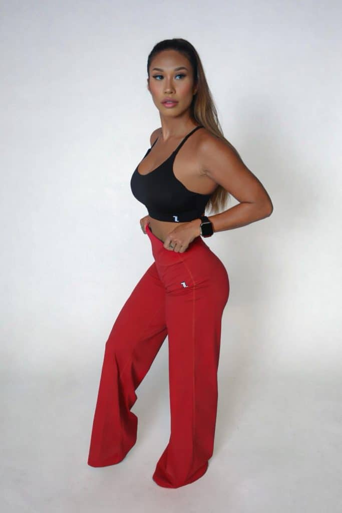 A woman poses against a plain white background, wearing a black sports bra, high-waisted red wide-leg pants, and a smartwatch. She stands sideways, looking ahead with one knee bent and hands resting on her thighs.
