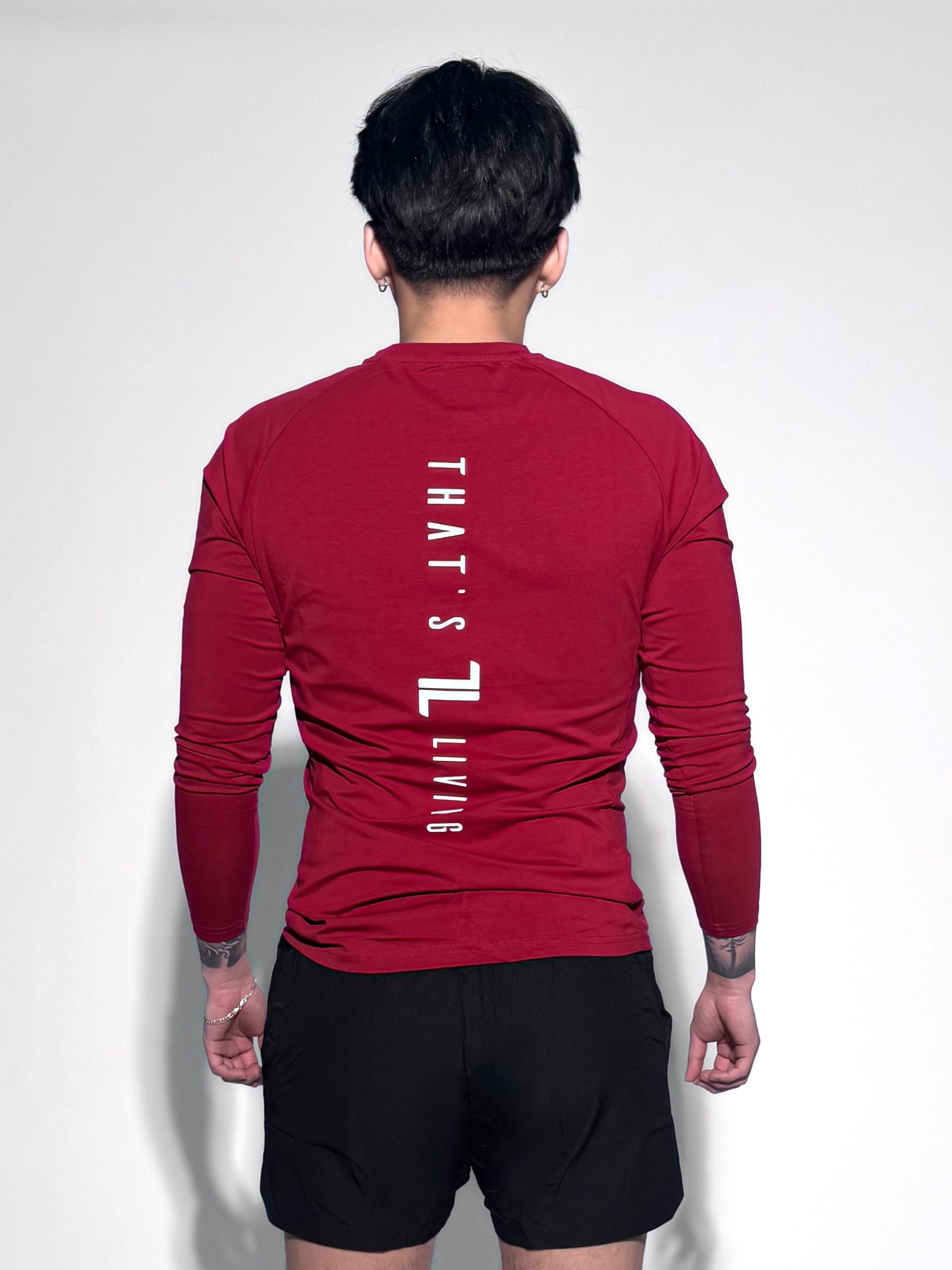 A person wearing a long-sleeve red shirt with the text "THAT'S LIVING" printed vertically on the back stands facing away, also wearing black shorts against a plain white background.