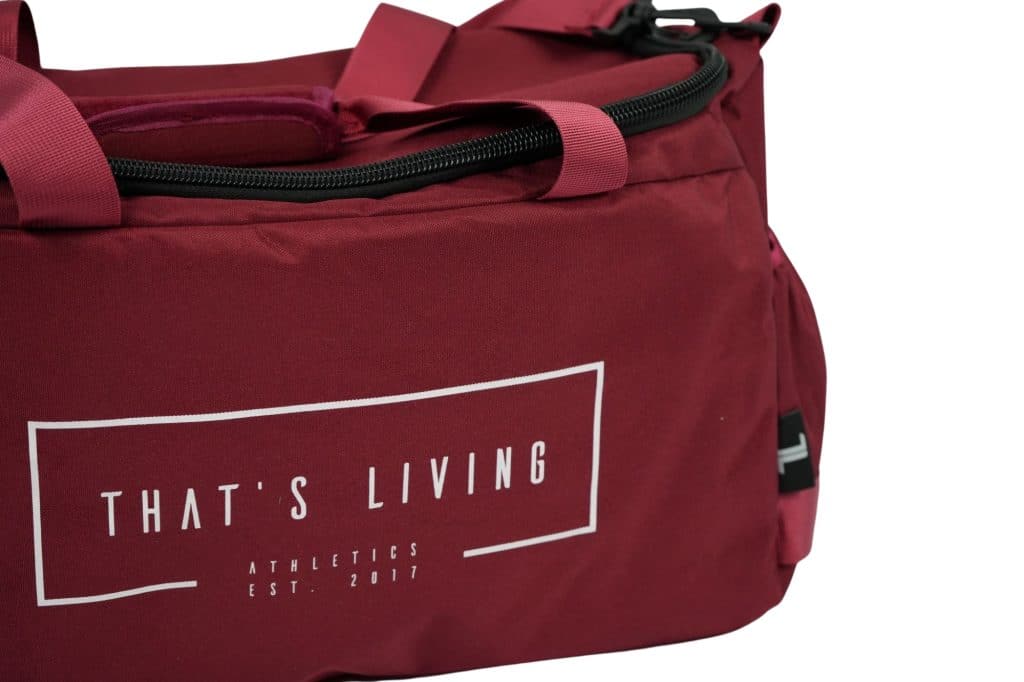 A close-up of a maroon duffel bag with the words "THAT'S LIVING ATHLETICS EST. 2017" printed in white on the side. The bag has black zippers and straps.