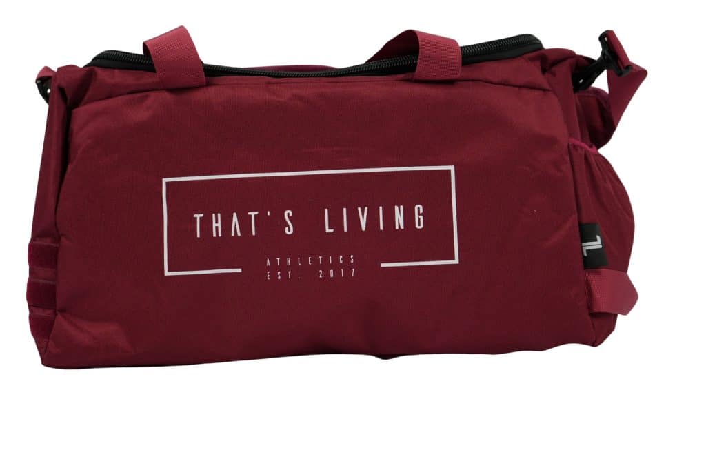 A red duffel bag with “THAT'S LIVING” and “ATHLETICS EST. 2017” printed in white on the side. The bag has short handles and a zipper on top.