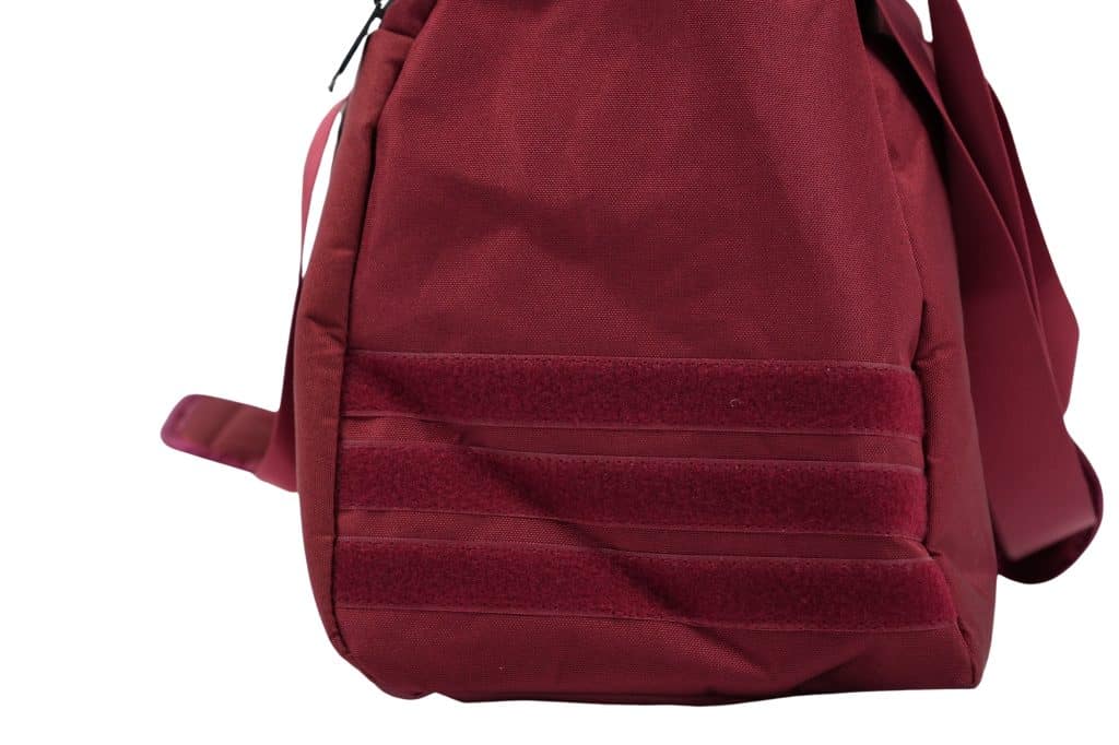 A close-up of the side of a red TRIARC Duffle Bag, featuring three horizontal Velcro strips and a shoulder strap visible on the left side.