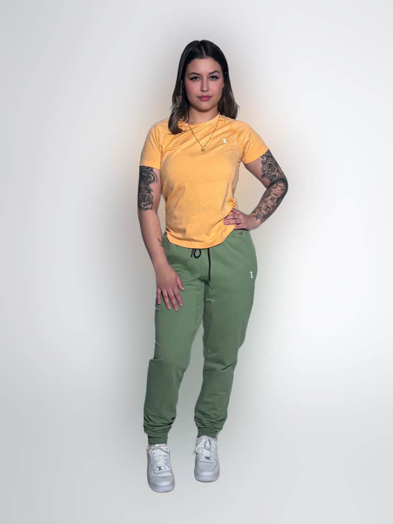 A woman with long dark hair wearing a yellow T-shirt, green jogger pants, and white sneakers stands confidently against a plain white background. She has tattoos on both arms and is looking at the camera with a neutral expression.
