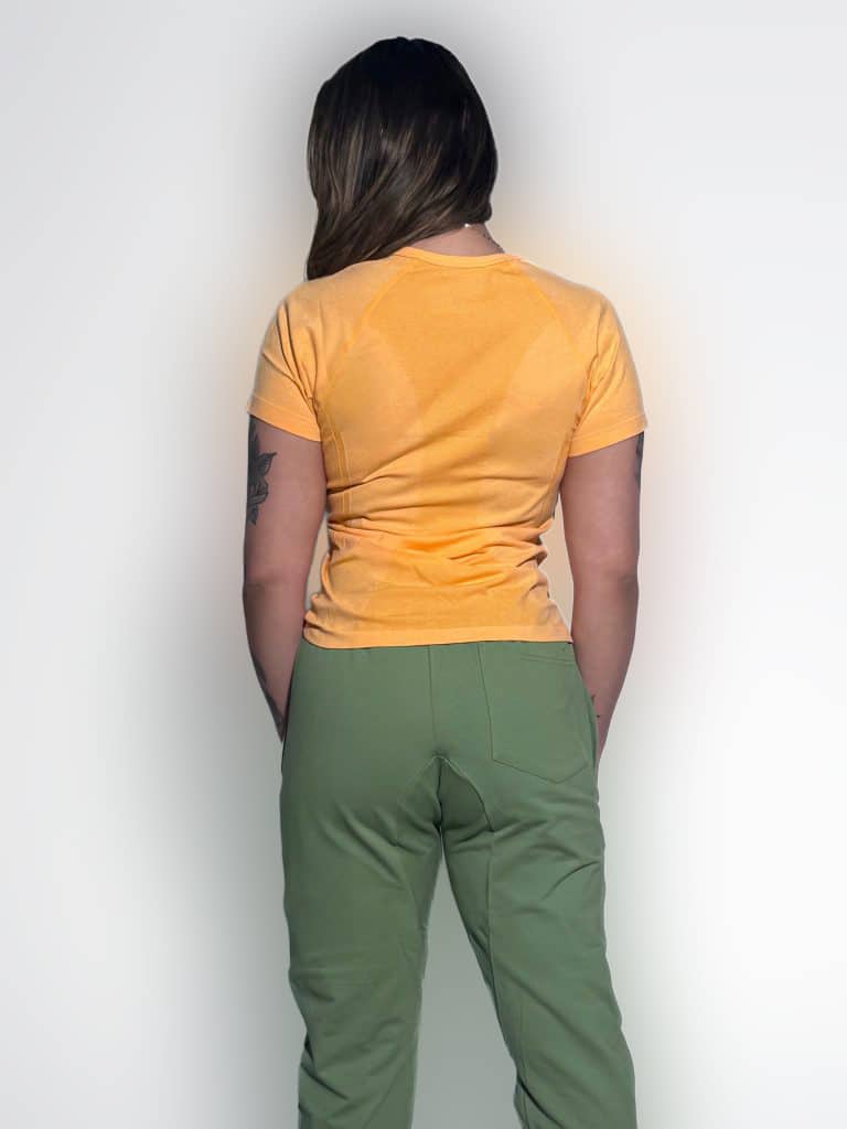 A person with long brown hair stands facing away, wearing a short-sleeved orange shirt and green pants. A tattoo is visible on their left upper arm. The background is plain white.