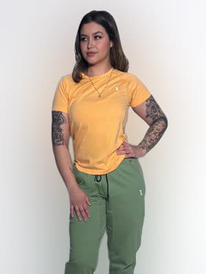 A woman with tattoos on her arms stands against a plain white background. She is wearing a yellow T-shirt, green pants, and gold necklaces, with her right hand on her hip and her left arm relaxed.
