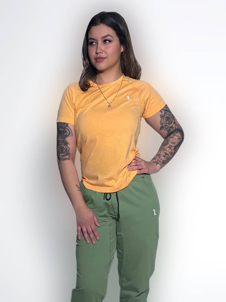 A woman with tattoos on her arms stands against a plain white background. She is wearing a yellow T-shirt, green pants, and gold necklaces, with her right hand on her hip and her left arm relaxed.