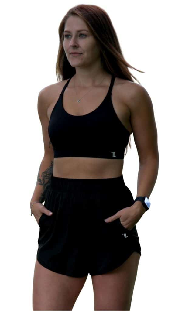 A woman with long brown hair stands with her hands in the pockets of her black athletic shorts, wearing a black sports bra, a smartwatch, and a necklace. She has a tattoo on her left arm and is looking slightly to the side.