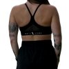 A woman with long brown hair and tattoos stands with her back to the camera, wearing a black Everyday Ease Bra featuring white text on the back and paired with black athletic shorts.