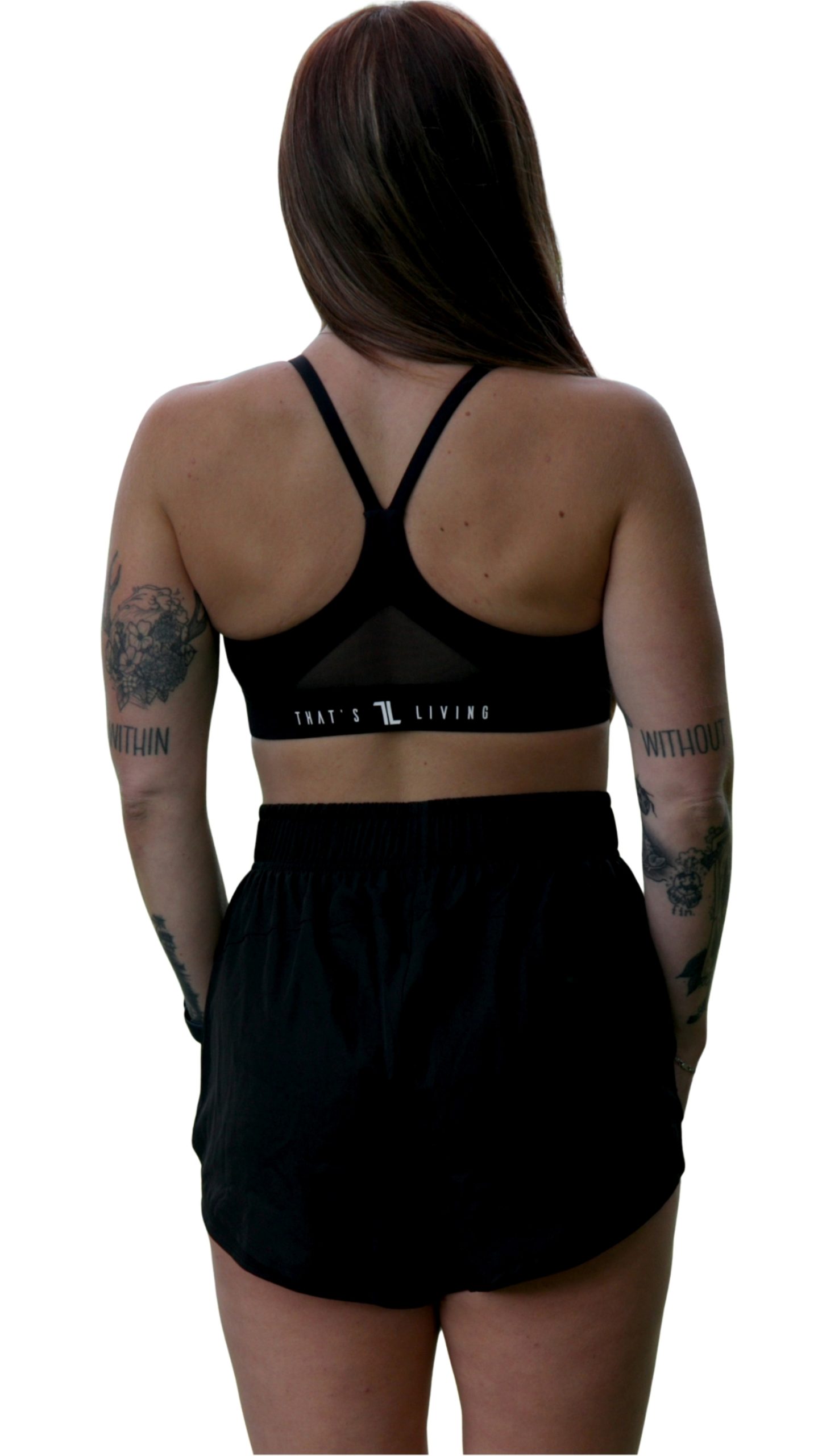 A woman with long brown hair and tattoos stands with her back to the camera, wearing a black Everyday Ease Bra featuring white text on the back and paired with black athletic shorts.