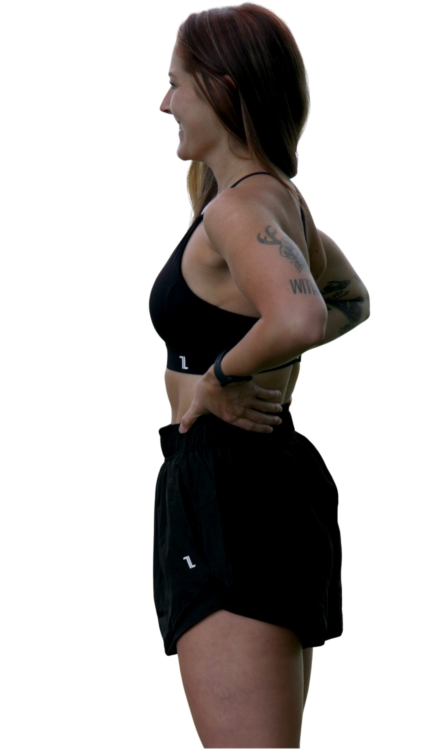 A woman with long brown hair stands in profile, smiling and wearing the black Everyday Ease Bra and black shorts. She has tattoos on her arm and her hands are on her hips.