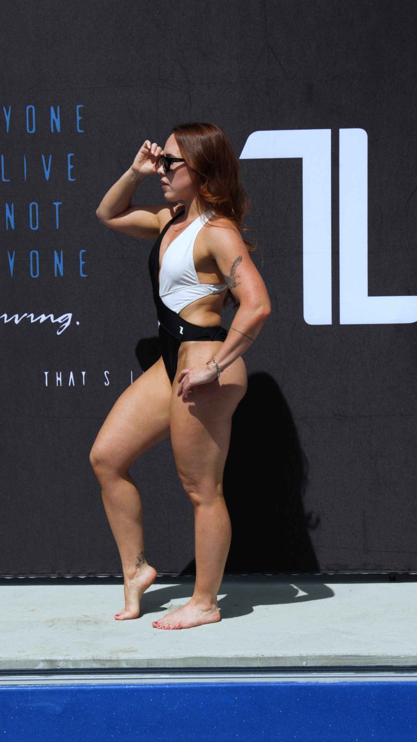 A woman wearing the Womens Vita One Swim Suit stands barefoot by a pool, posing with one hand on her sunglasses and the other on her hip, in front of a black backdrop with white text.
