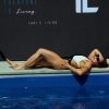 Wearing the Womens Vita One Swim Suit, a woman lounges by a pool in the sun. Behind her, a dark wall shows the text: "Everyone is alive but not everyone is living. That's living.
