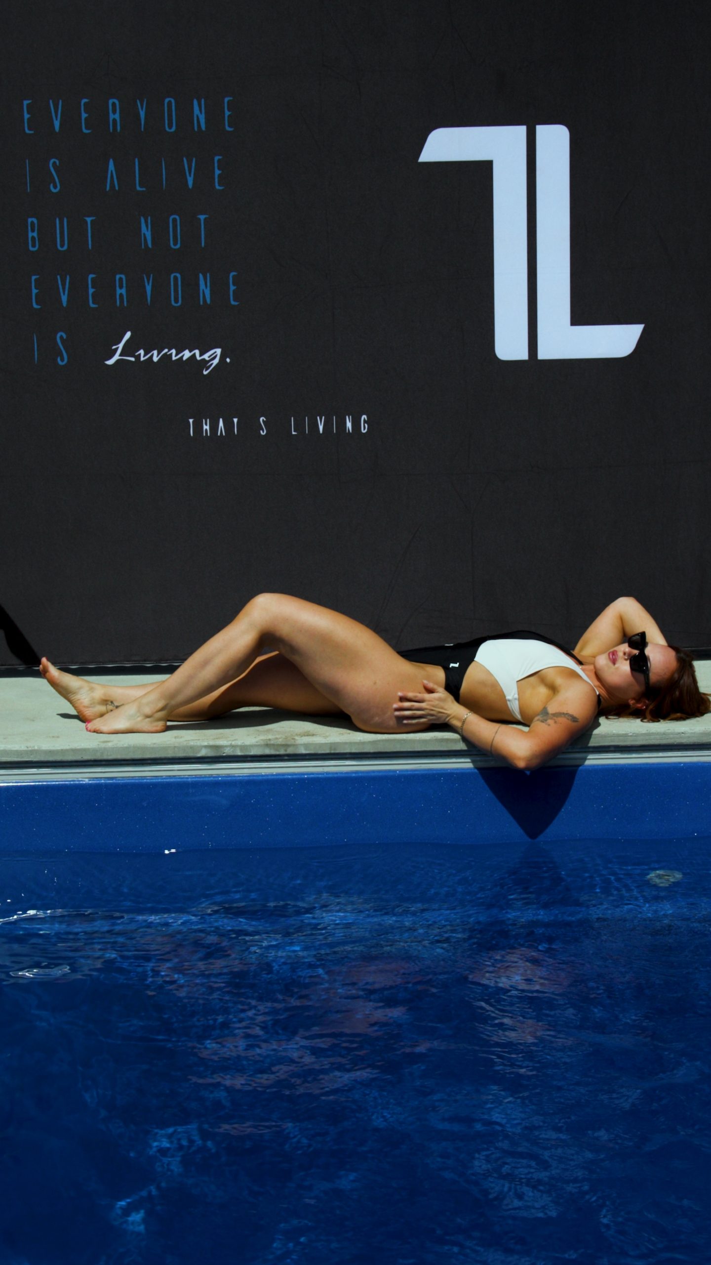 Wearing the Womens Vita One Swim Suit, a woman lounges by a pool in the sun. Behind her, a dark wall shows the text: "Everyone is alive but not everyone is living. That's living.