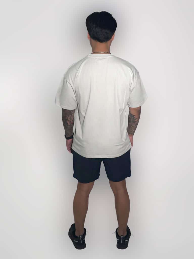 A person with short dark hair stands facing away, wearing a white t-shirt, black shorts, and black sneakers. Their arms are by their sides, and they have tattoos on both arms. The background is plain and white.