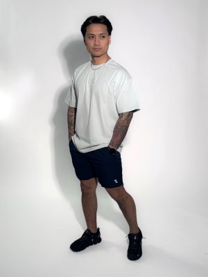 A man with tattoos poses against a white background, wearing a loose white T-shirt, dark athletic shorts, black sneakers, and a necklace, with hands in pockets and looking slightly to the side.