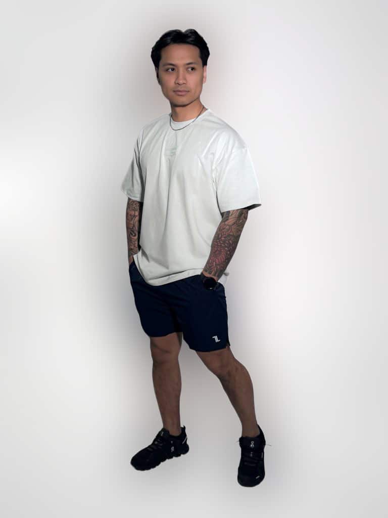 A man with tattoos poses against a white background, wearing a loose white T-shirt, dark athletic shorts, black sneakers, and a necklace, with hands in pockets and looking slightly to the side.